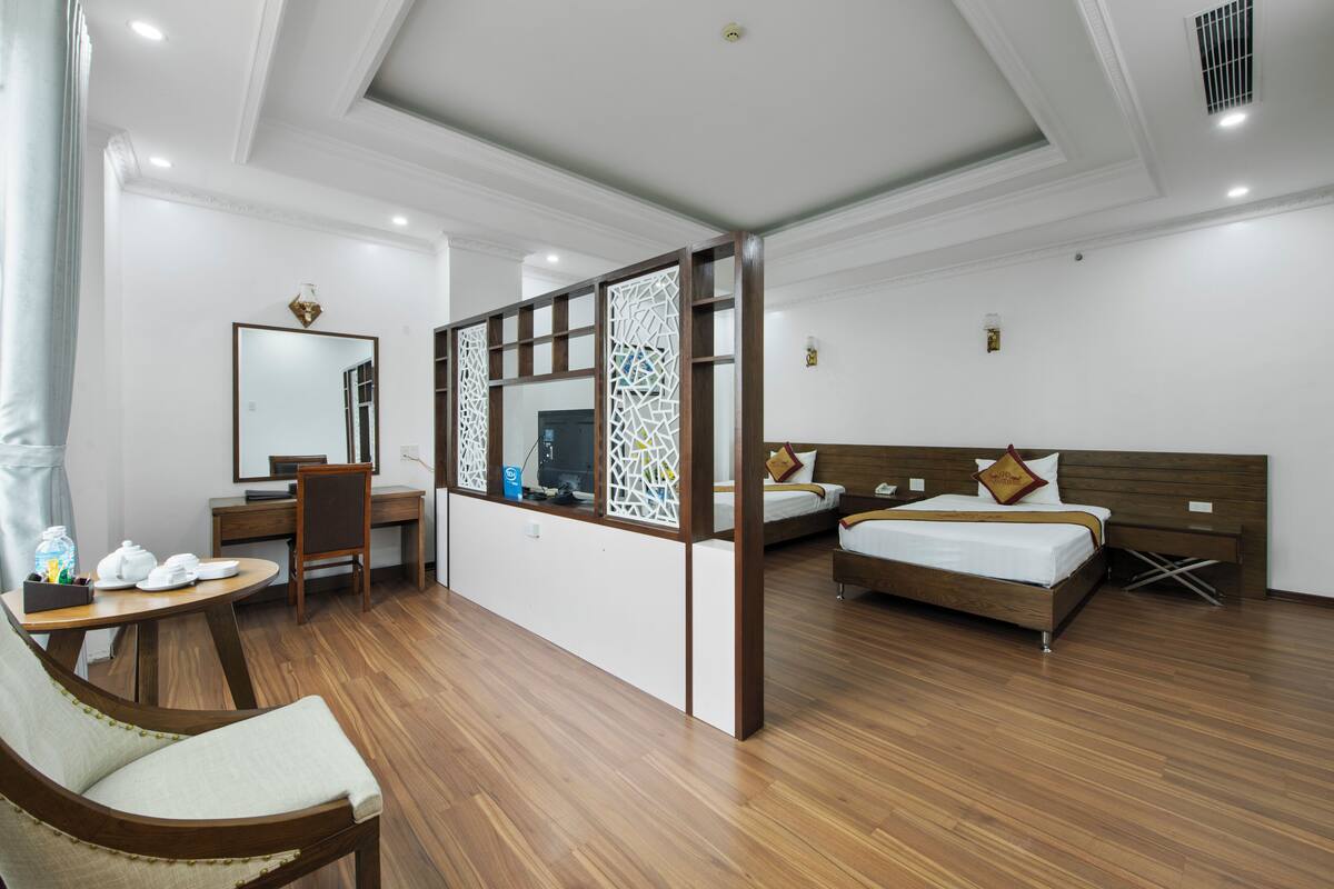 A hotel room with a wooden floor, a desk with a chair, a bed with a wooden headboard, a nightstand, a mirror, and a decorative screen.