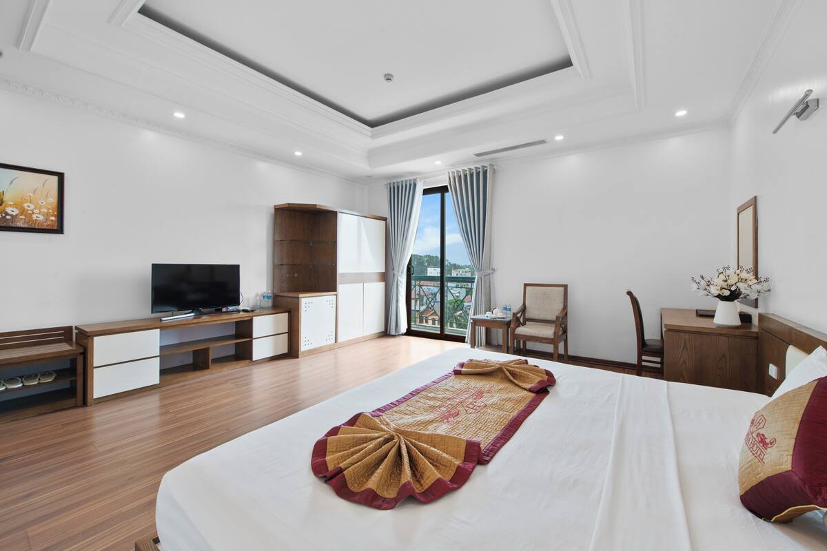 A hotel room with a large bed, a TV, a desk with a chair, and a window with curtains.