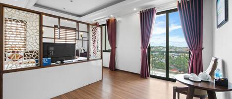 A modern hotel room with a wooden floor, a flat-screen TV, a small table with a teapot and cups, and a large window with curtains.