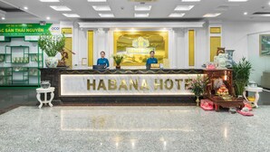 A hotel reception desk with a sign reading "HABANA HOTEL". There are two receptionists seated behind the desk. The reception area has a marble floor and is decorated with plants and a small statue.