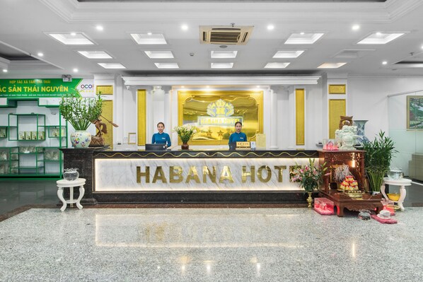 Un mostrador de recepción de hotel con un letrero que dice "HABANA HOTEL". Dos recepcionistas están sentadas detrás del mostrador. La zona de recepción tiene un piso de mármol y está decorada con plantas y una pequeña estatua.