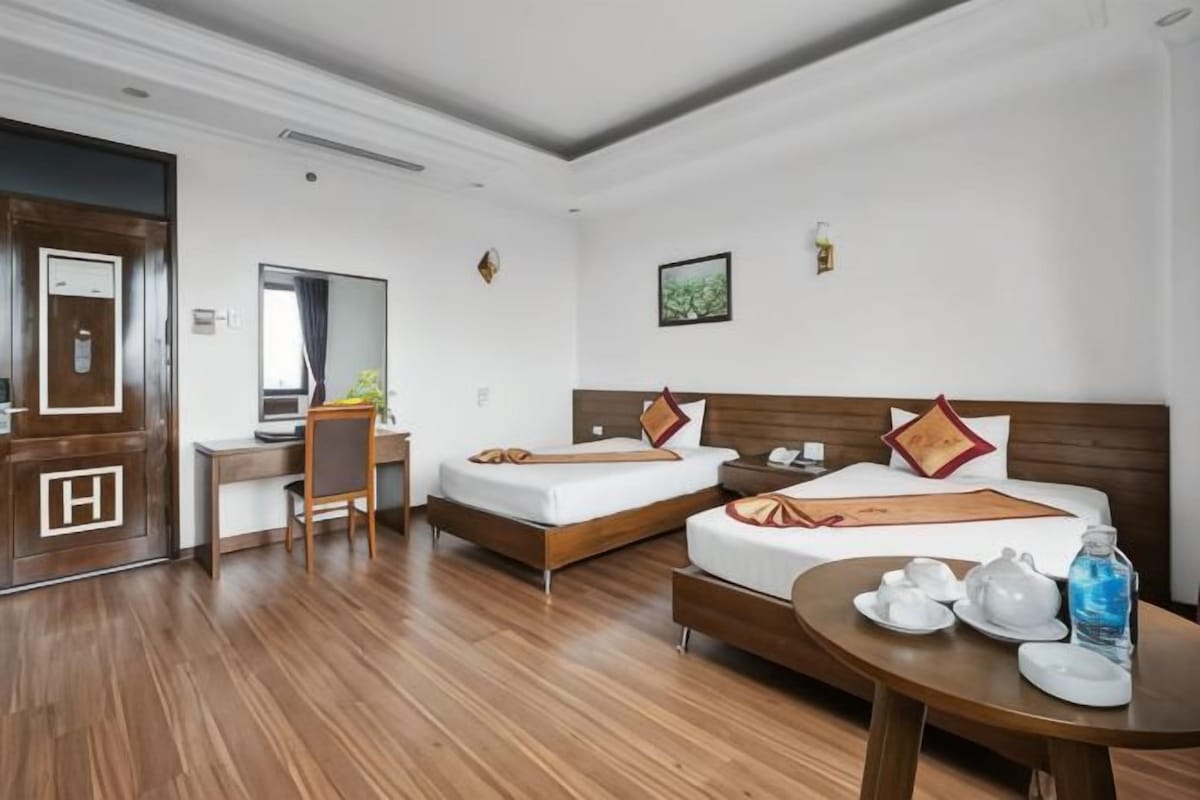 Habitació d'hotel amb dos llits, un escriptori, una cadira, una taula rodona amb servei de te i una porta amb el símbol 'H'.