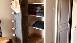 Una habitación con piso de madera, una silla, un banco, una puerta y un estante con mantas dobladas.