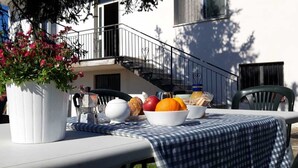 A table set outdoors with a white tablecloth, a white vase with pink flowers, a coffee pot, a teapot, a bowl of fruit, and a bottle of juice.