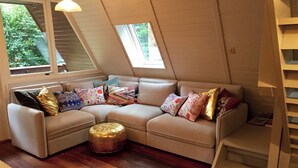 Un salón acogedor con un sofá modular, un taburete redondo y suelo de madera. Dos ventanas dejan entrar luz natural y ofrecen vistas al verdor exterior.