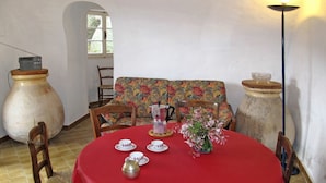 Uma sala de jantar com uma mesa redonda coberta por uma toalha vermelha, rodeada por cadeiras de madeira. Há dois grandes vasos de cerâmica em cada lado da mesa, um arranjo floral num vaso sobre a mesa, e uma cafeteira na mesa.