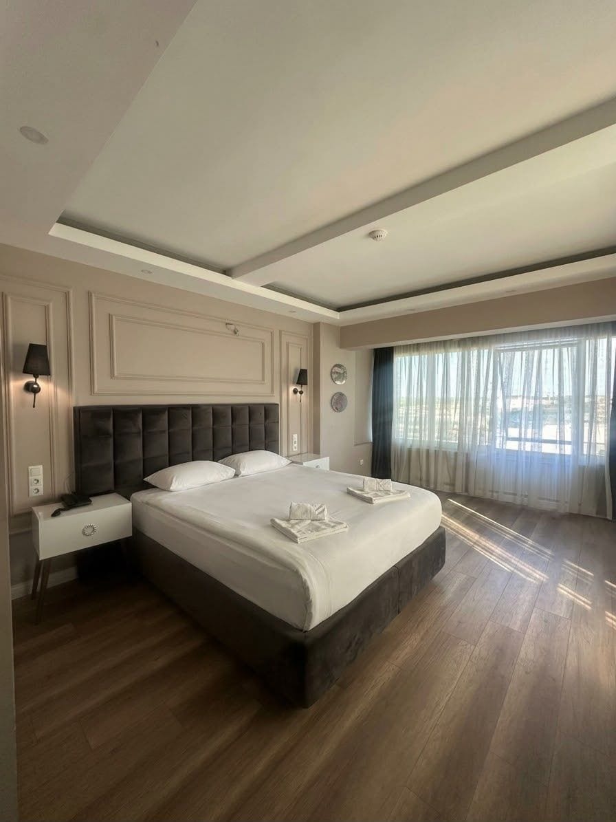 A modern hotel room with a large bed, bedside tables, and a view of the outdoors through sheer curtains.