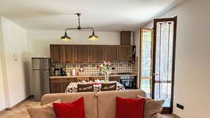 Un salón acogedor con un sofá, cojines rojos y un plaid estampado. Se ve una cocina con armarios de madera y un salpicadero de azulejos al fondo.