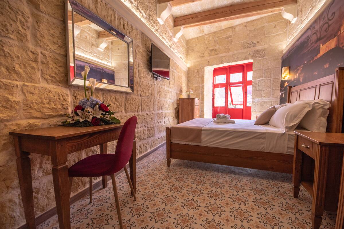 Une chambre avec un mur en pierre, un lit en bois, un bureau avec une chaise et une fenêtre rouge.