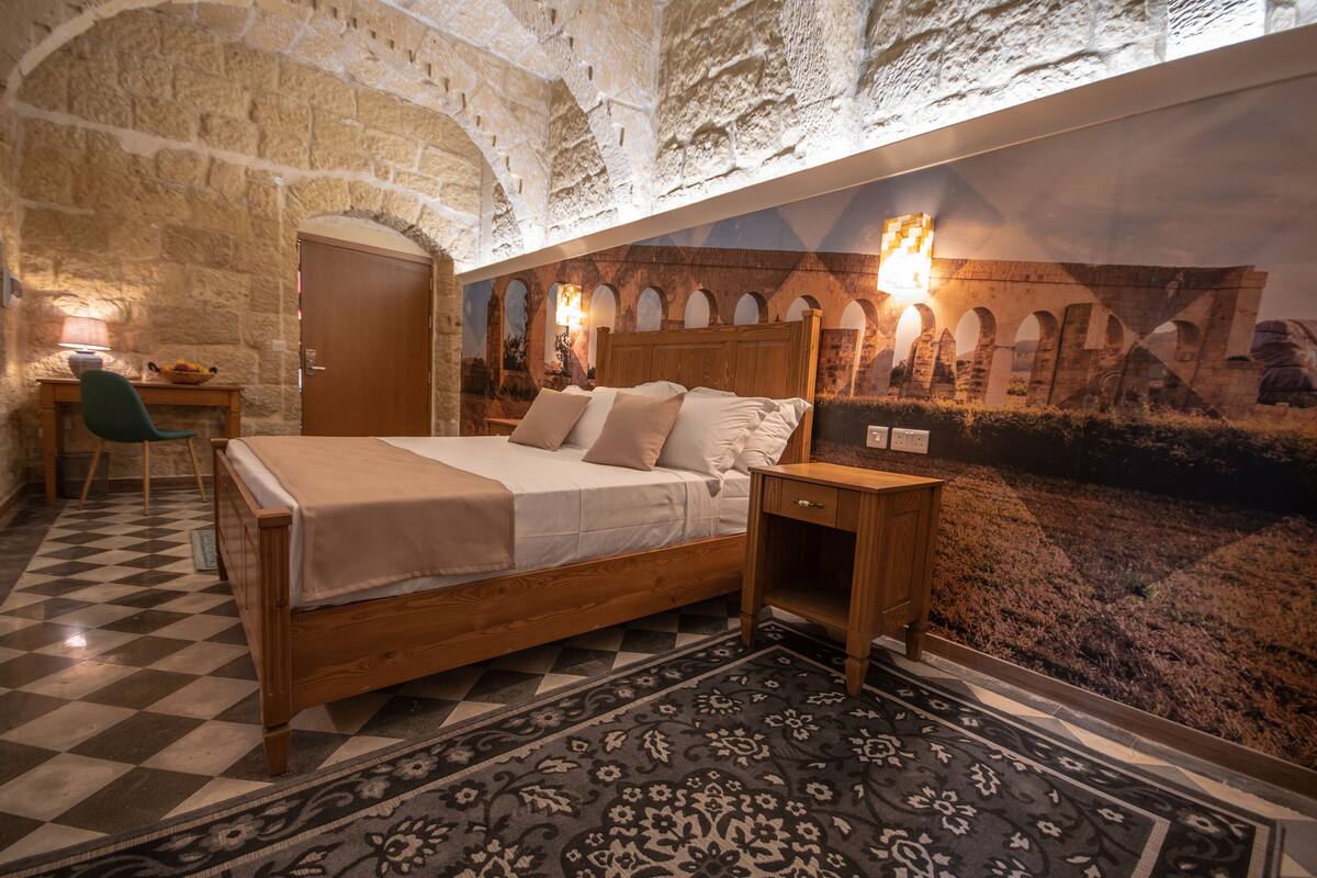 Une chambre avec un sol à carreaux, un lit en bois, une table de chevet, un bureau et une fresque murale représentant une structure ancienne.