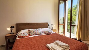 Ein ordentlich gemachtes Bett mit roter Decke, zwei Kissen mit bunten Mustern und einem Satz gefalteter Handtücher auf dem Boden.