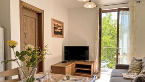 Ein Essbereich mit Holztisch, Stühlen, einer Vase mit Blumen, einem Fernseher und einem Balkon mit Blick auf Grünflächen.