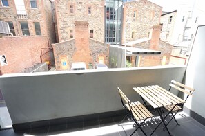 A balcony with a table and chairs, overlooking a courtyard with brick buildings and a modern glass structure. - East End CBD Apt-2 Queen beds+sofa bed+smart tv (Adelaide)