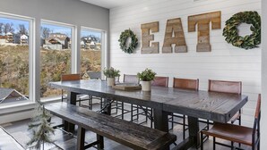 A dining area with a large wooden table and benches, a view of houses through large windows, and a wall with wooden letters spelling "EAT" and a wreath.