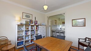 Un comedor con mesa y sillas de madera, una estantería, y una cocina visible a través de un vano abierto.
