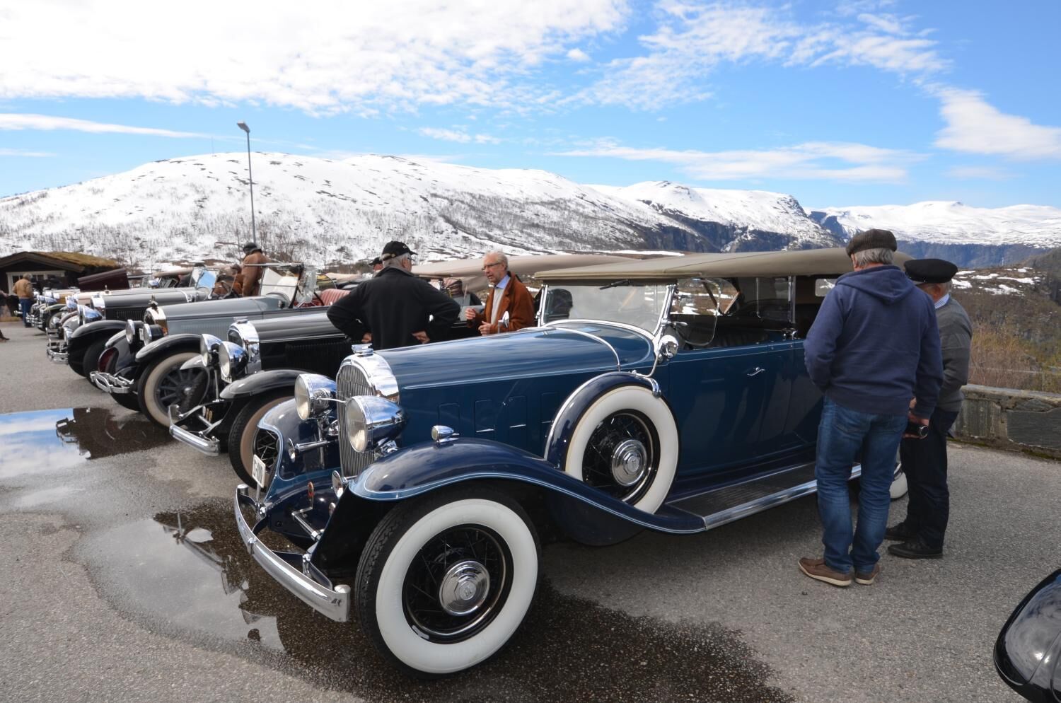 A vintage car exhibition with classic cars parked in a row, set against a snowy mountain backdrop.