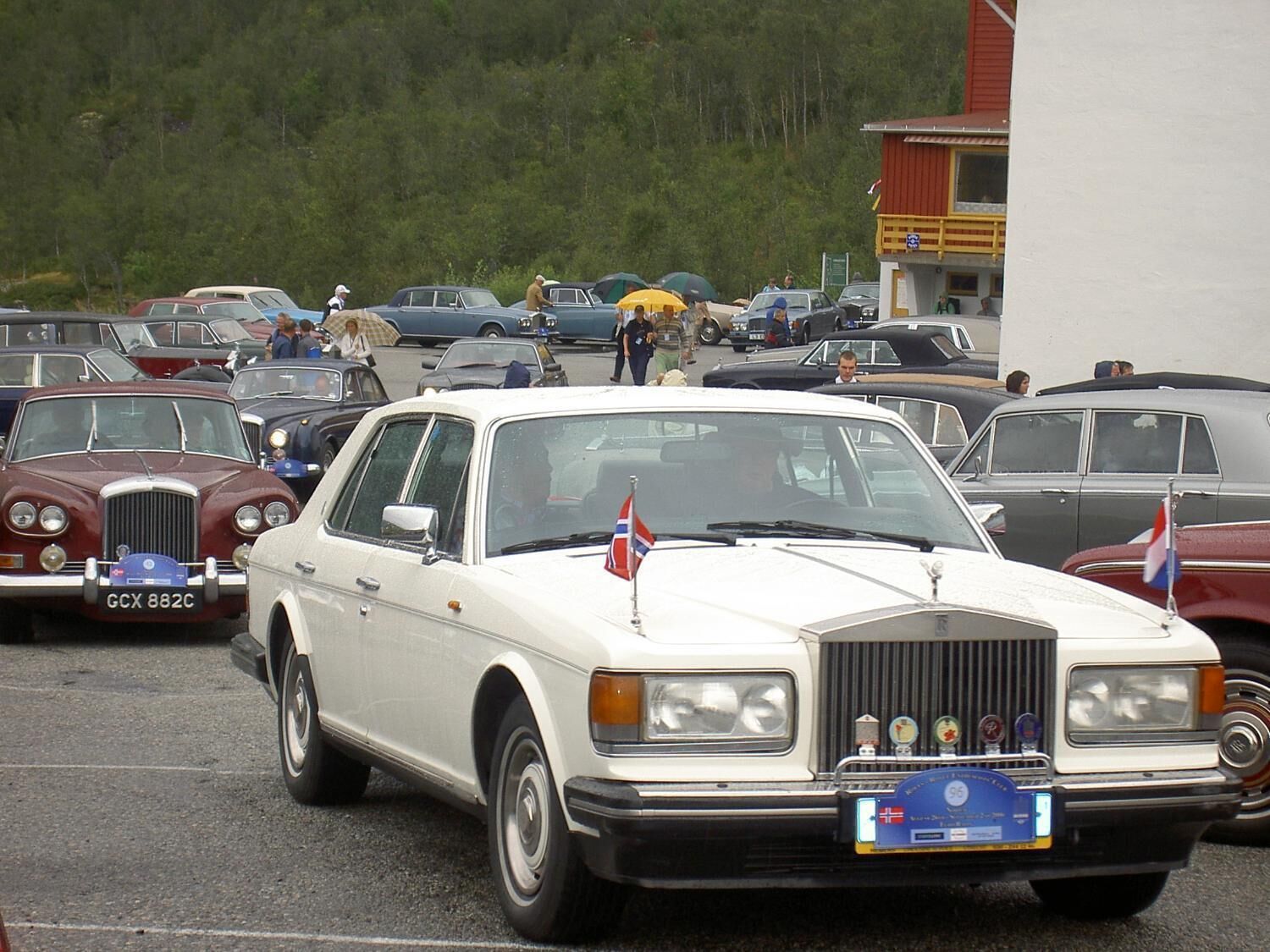 Un coche antiguo con una bandera, estacionado en un estacionamiento lleno de otros vehículos clásicos.