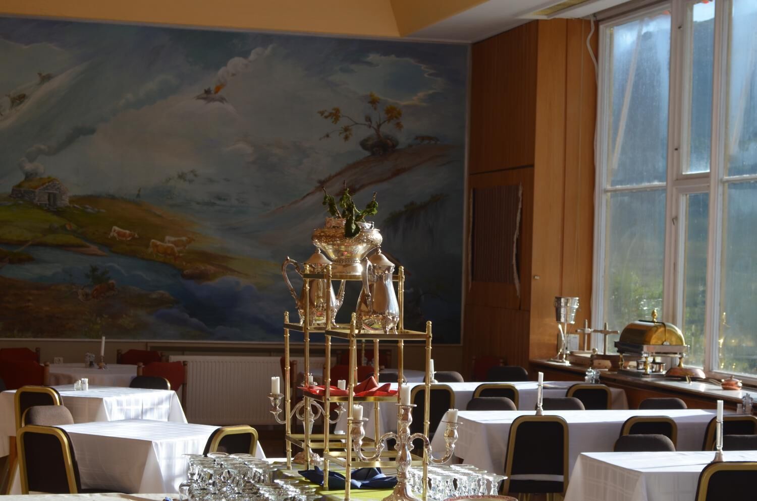 Un espace repas avec des tables dressées pour le service, une grande fresque murale et un présentoir décoratif en argent placé au centre.