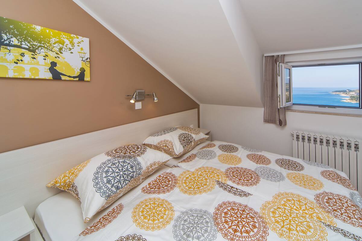 A bedroom with a bed, a nightstand, a wall-mounted lamp, a window with a view of the sea, and a painting on the wall.