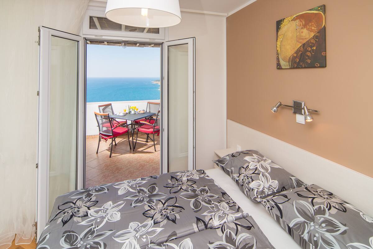 A bedroom with a bed, a view of the sea, and a table with chairs outside.