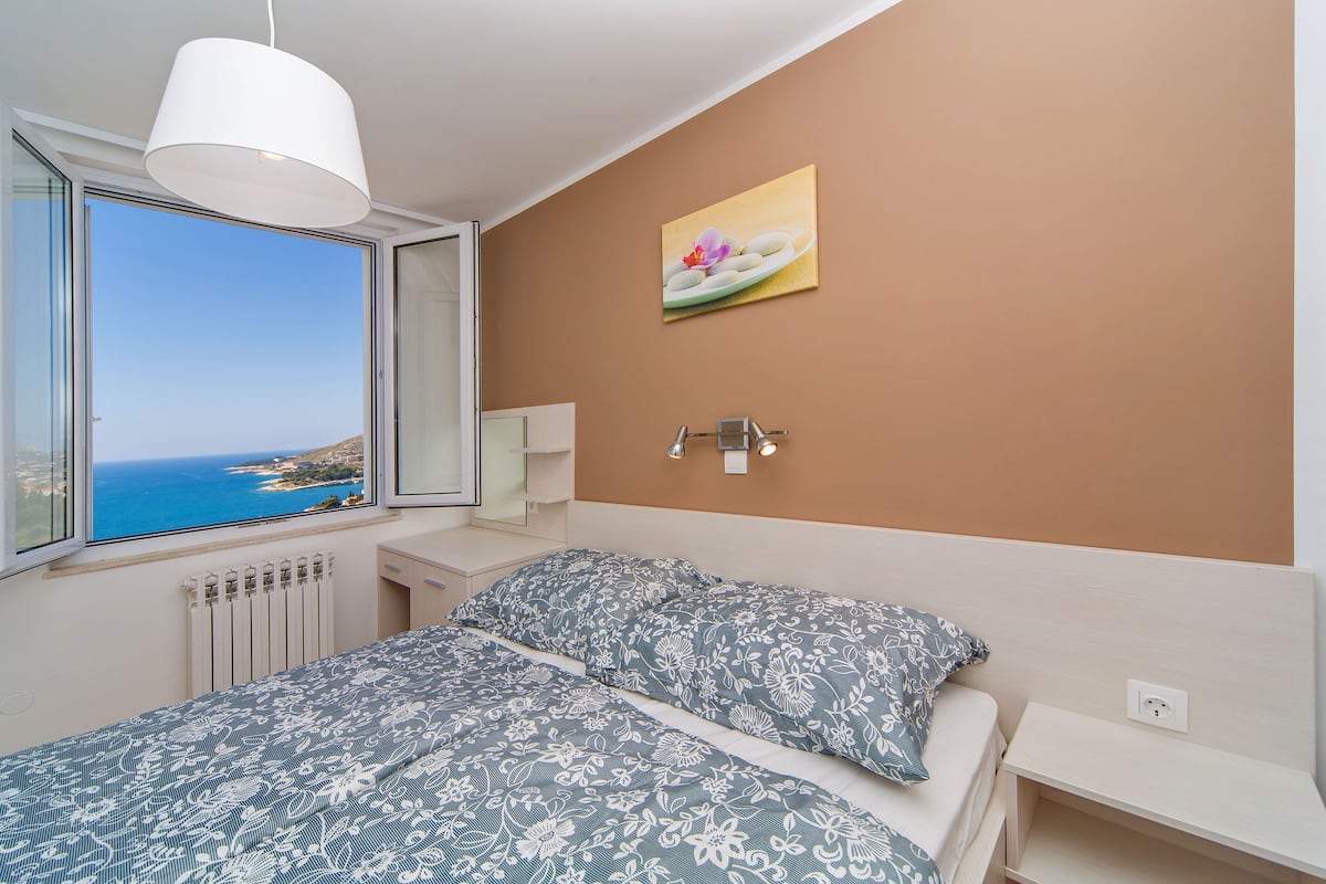A bedroom with a bed, a window with a view of the sea, a painting on the wall, and a bedside lamp.