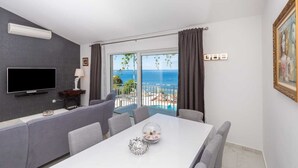 A modern living room with a flat-screen TV, grey sofa, dining table, and a view of the sea through large windows. - Villa Laura (Zupa dubrovacka)