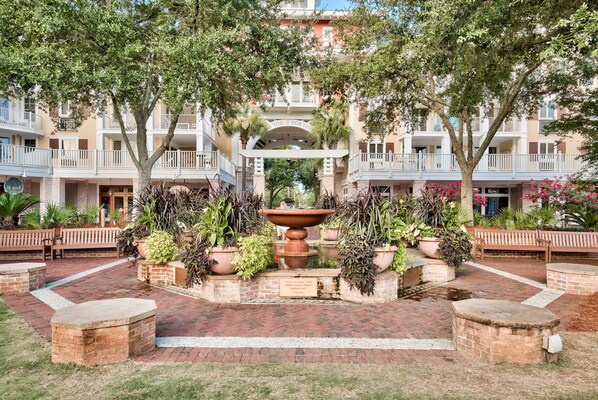 A courtyard with a central fountain, surrounded by potted plants and benches, with a building featuring balconies in the background.