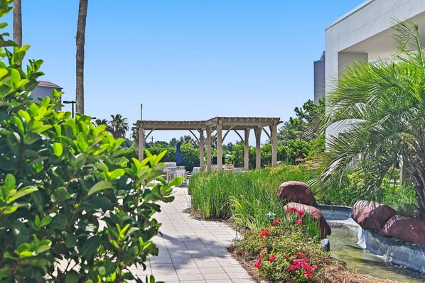 A garden pathway with a pergola, palm trees, and a water feature.