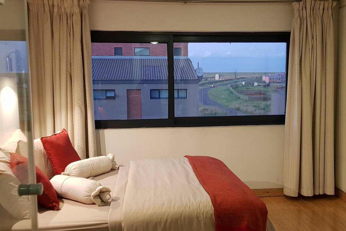 A hotel room with a large window, a bed with red and white bedding, and a view of buildings and greenery outside.
