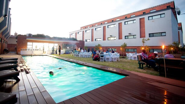 A swimming pool with wooden deck seating, surrounded by a building with multiple windows and a brick facade. - Inn Green at The Lofts (Secunda)