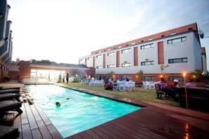 Una piscina con mobiliario de deck de madera, rodeada por un edificio de fachada de ladrillo y múltiples ventanas.