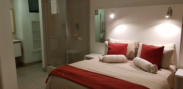 Un dormitorio moderno con cabina de ducha de vidrio, cama con ropa de cama blanca y almohadas rojas, y una mesita de noche.