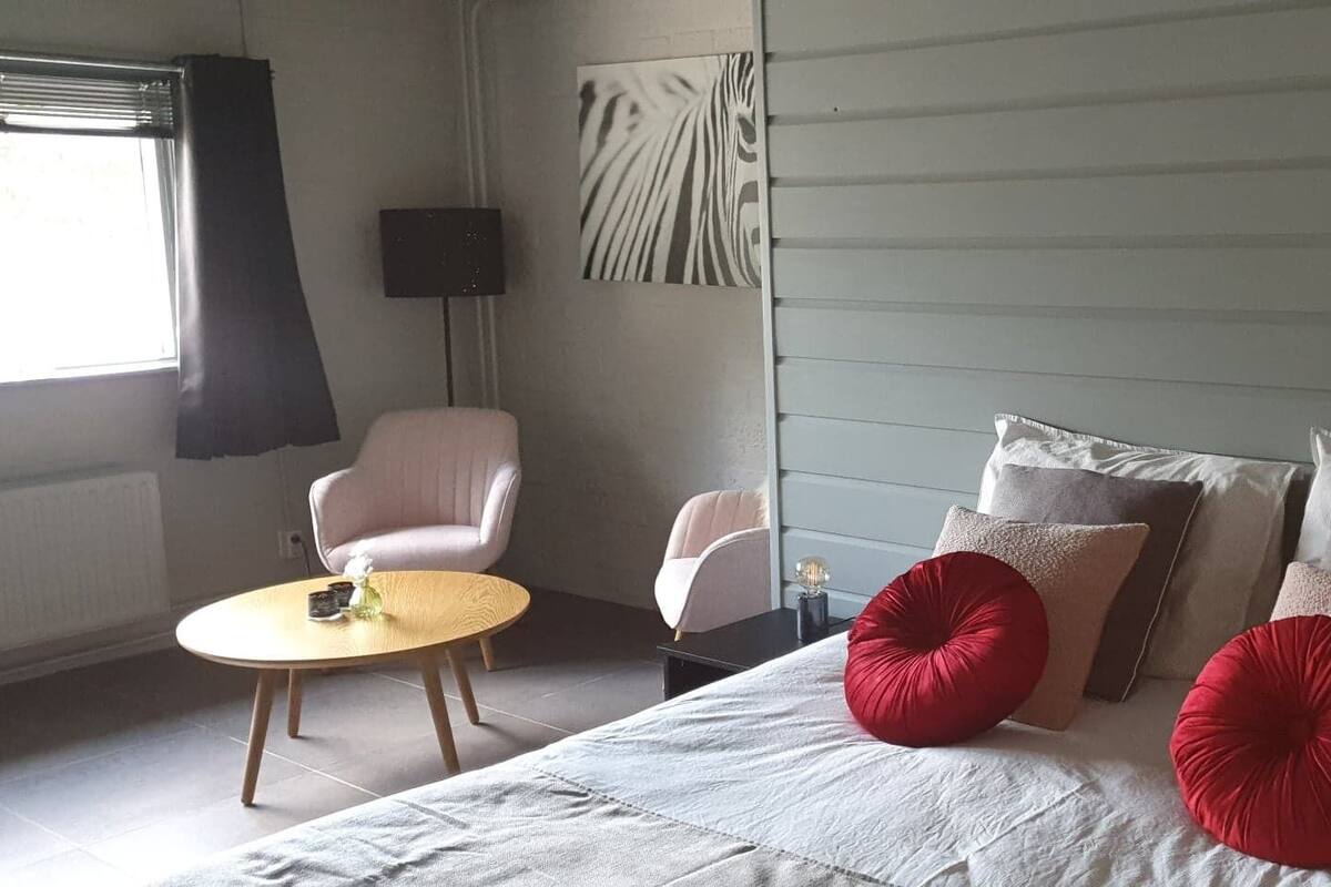 A bedroom with a bed, two chairs, a small table, and a wall-mounted light fixture.