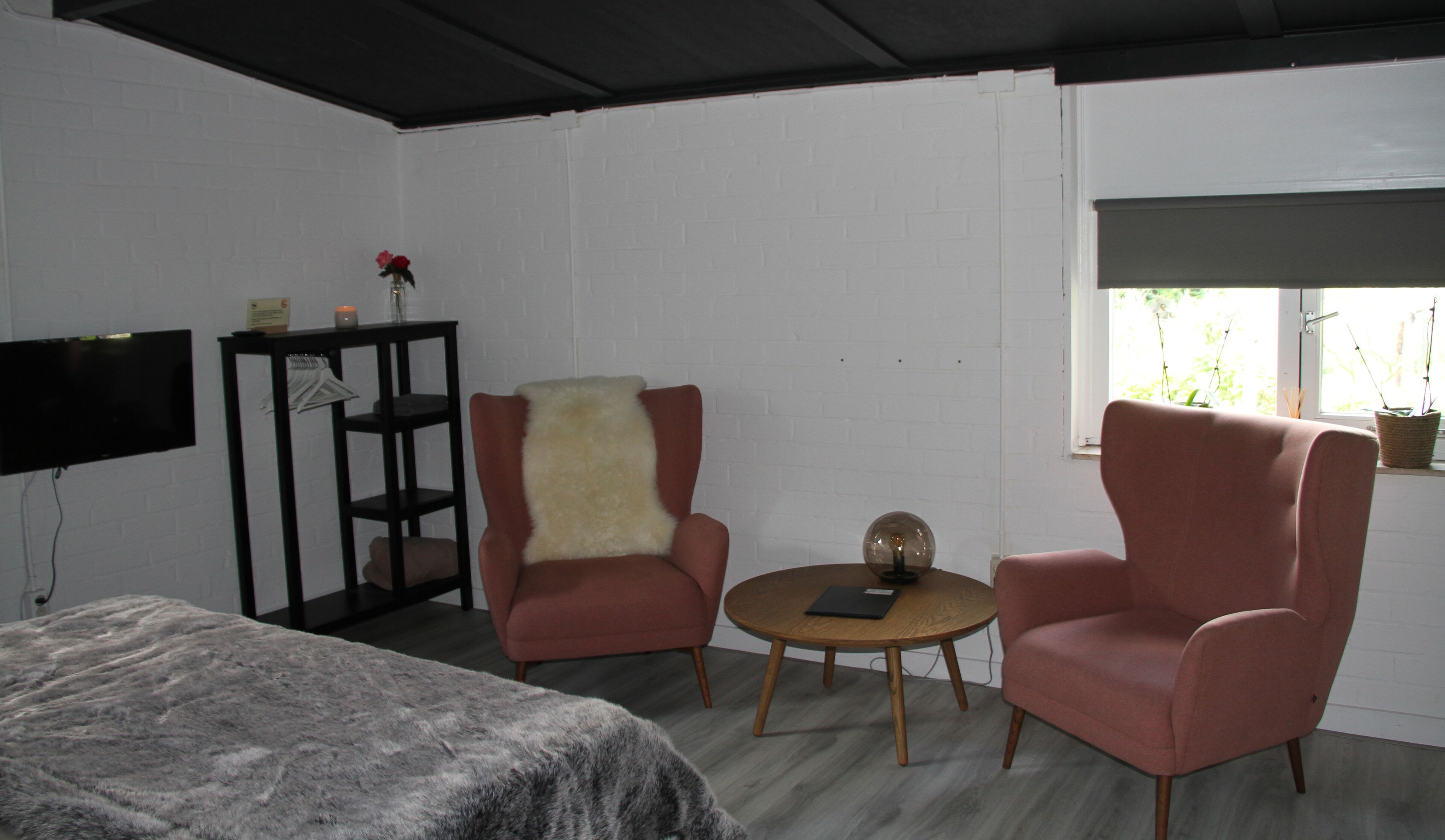 A room with a bed, two armchairs, a small round table, a TV on the wall, and a window with plants.