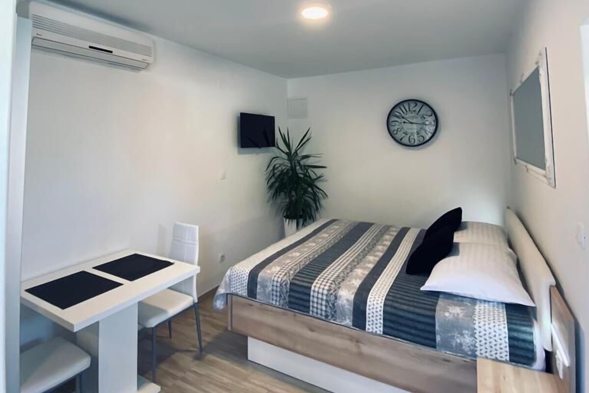 Une petite chambre à coucher moderne, avec un lit, un bureau et une chaise. Il y a une horloge au mur et une plante dans un coin.
