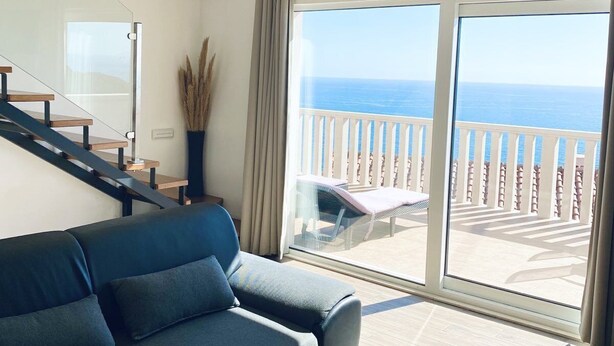 A modern living room with a sofa, a small table, and a view of the ocean through large sliding doors. - Marojica Apartments (Konavle)