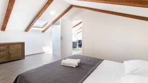 A bedroom with a sloped ceiling, wooden beams, a bed with white and grey bedding, a wooden wardrobe, and a view through an open door to another room. - Marojica Apartments (Konavle)