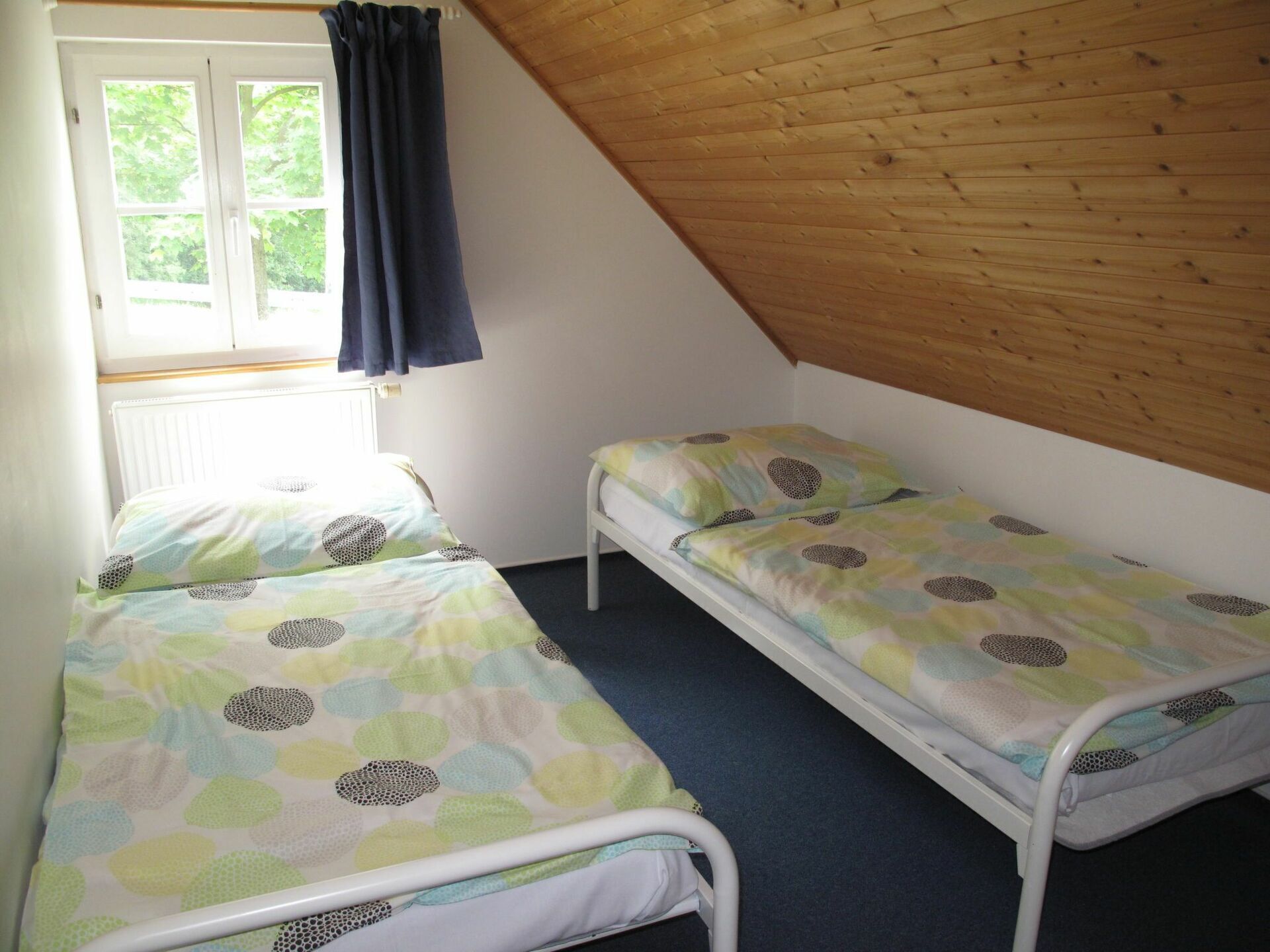 A small, single-room with two single beds, a window with curtains, and a sloped wooden ceiling.