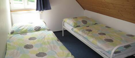 A small, single-room with two single beds, a window with curtains, and a sloped wooden ceiling.