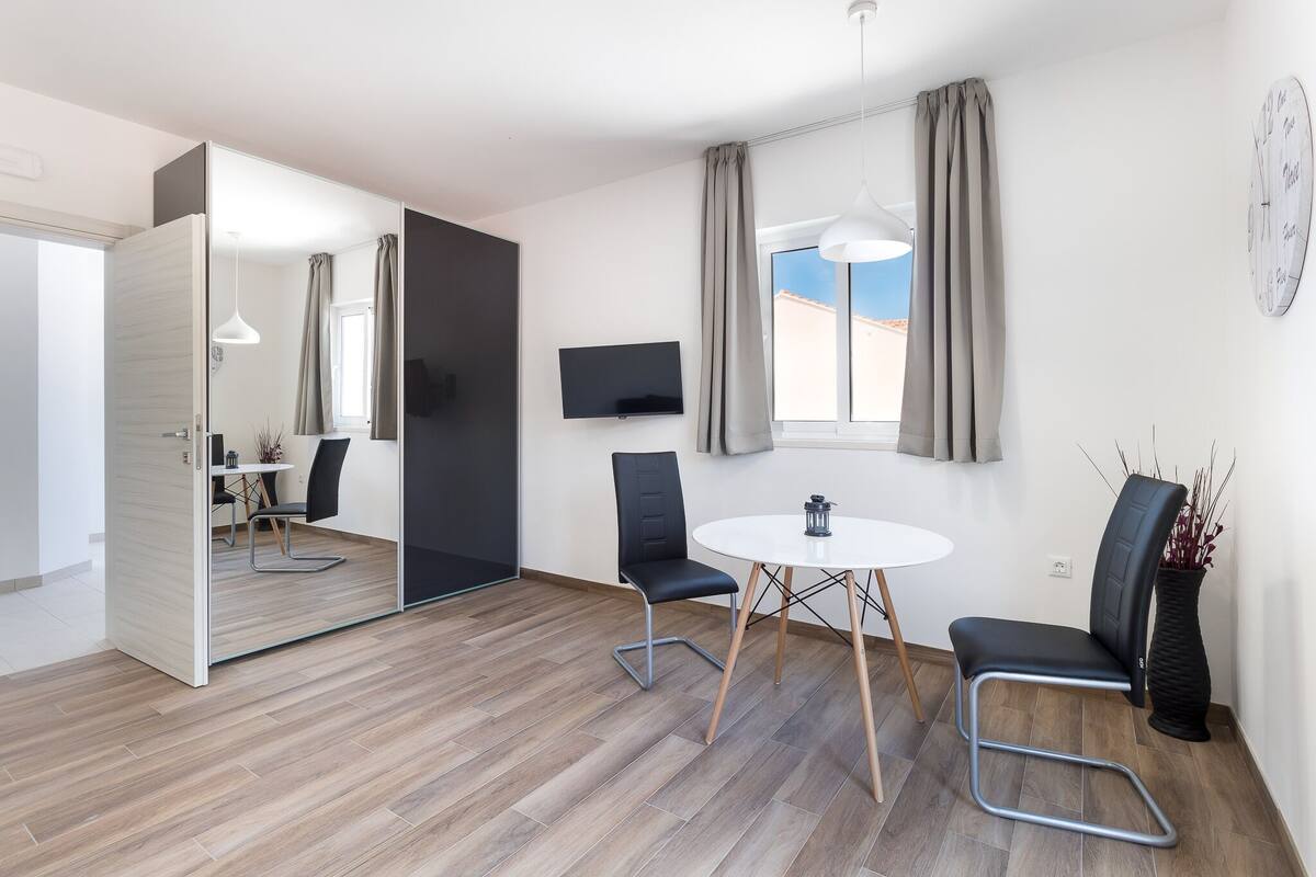 A modern room with a wooden floor, a round table, two black chairs, a TV on the wall, and a window with curtains.
