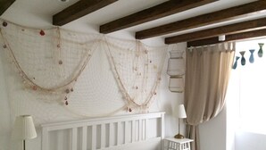 A bedroom with a white headboard, a bed with a grey blanket, a bedside table with a lamp, and a window with curtains. - Casa Karuza (Pag)