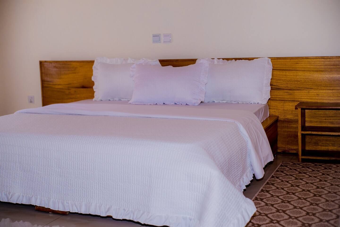A neatly made bed with white bedding and pillows, a wooden headboard, and a small wooden nightstand beside it.