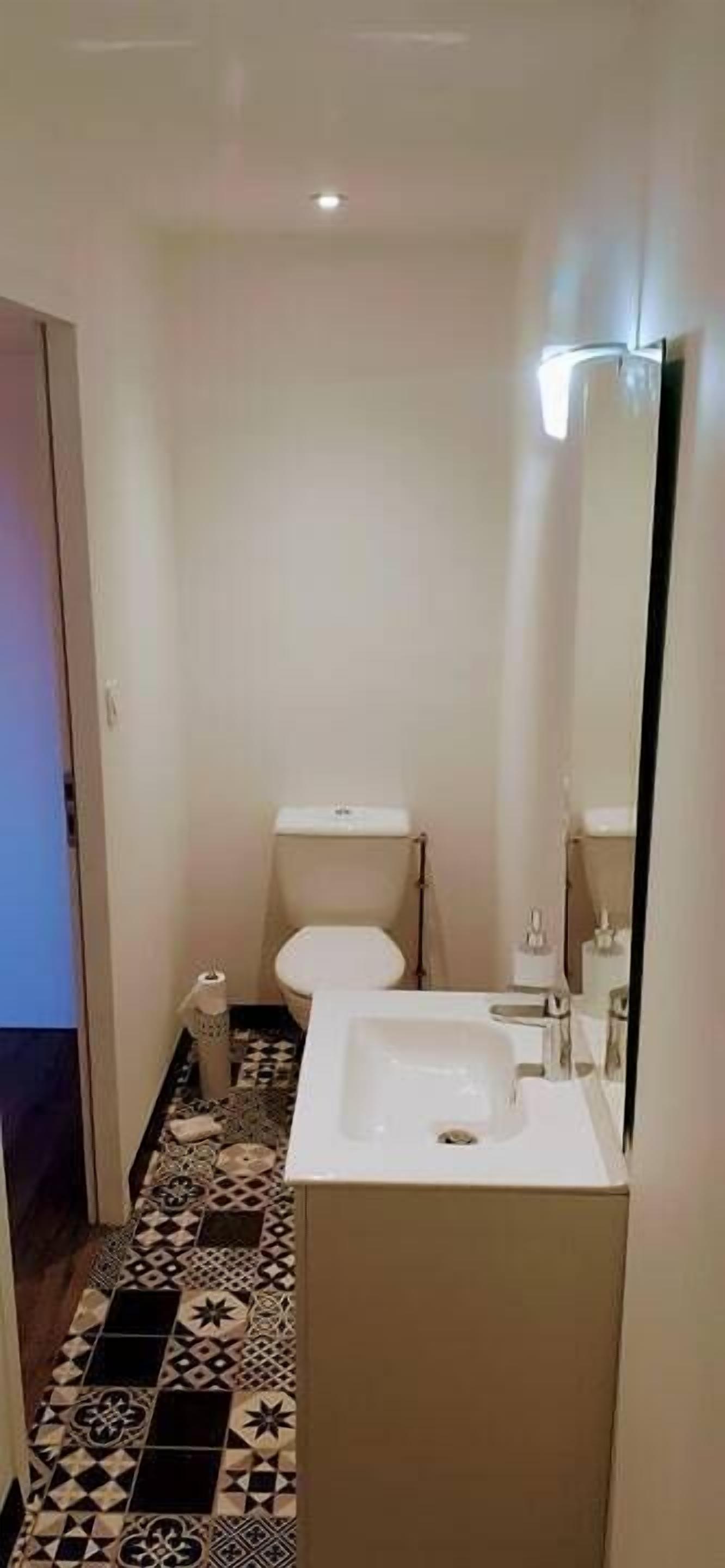 Een kleine badkamer met toilet, wastafel en spiegel. De vloer heeft een betegeld patroon.