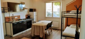 A compact kitchen with a stove, sink, and cabinets, a dining table with chairs, a bunk bed, and a window with a view of the street. - Résidences du Palmier (Saint-Laurent-sur-Sevre)