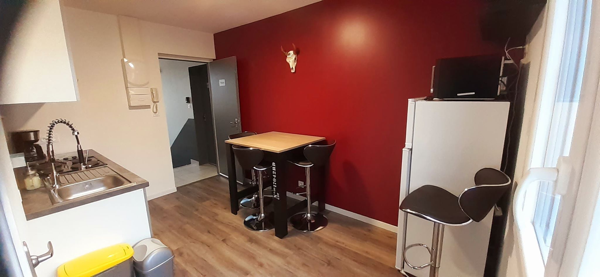 Eine kleine Küche mit Holzarbeitsplatte, Spüle und Kühlschrank. Eine rote Akzentwand und ein Essbereich mit Hocker sind vorhanden.