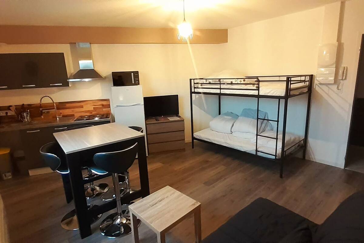 Un espace de vie compact comprenant un coin cuisine, un lit superposé, une table à manger avec des chaises et un petit canapé.