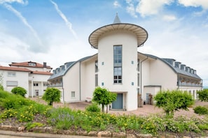 Un bâtiment blanc doté d’une tour centrale, entouré d’un jardin où poussent différentes plantes et fleurs.