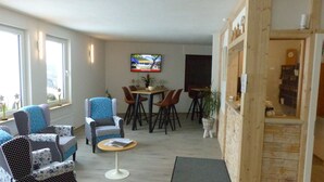 Un saló amb zona de menjador, televisió penjada a la paret i un moble de fusta amb rellotge.