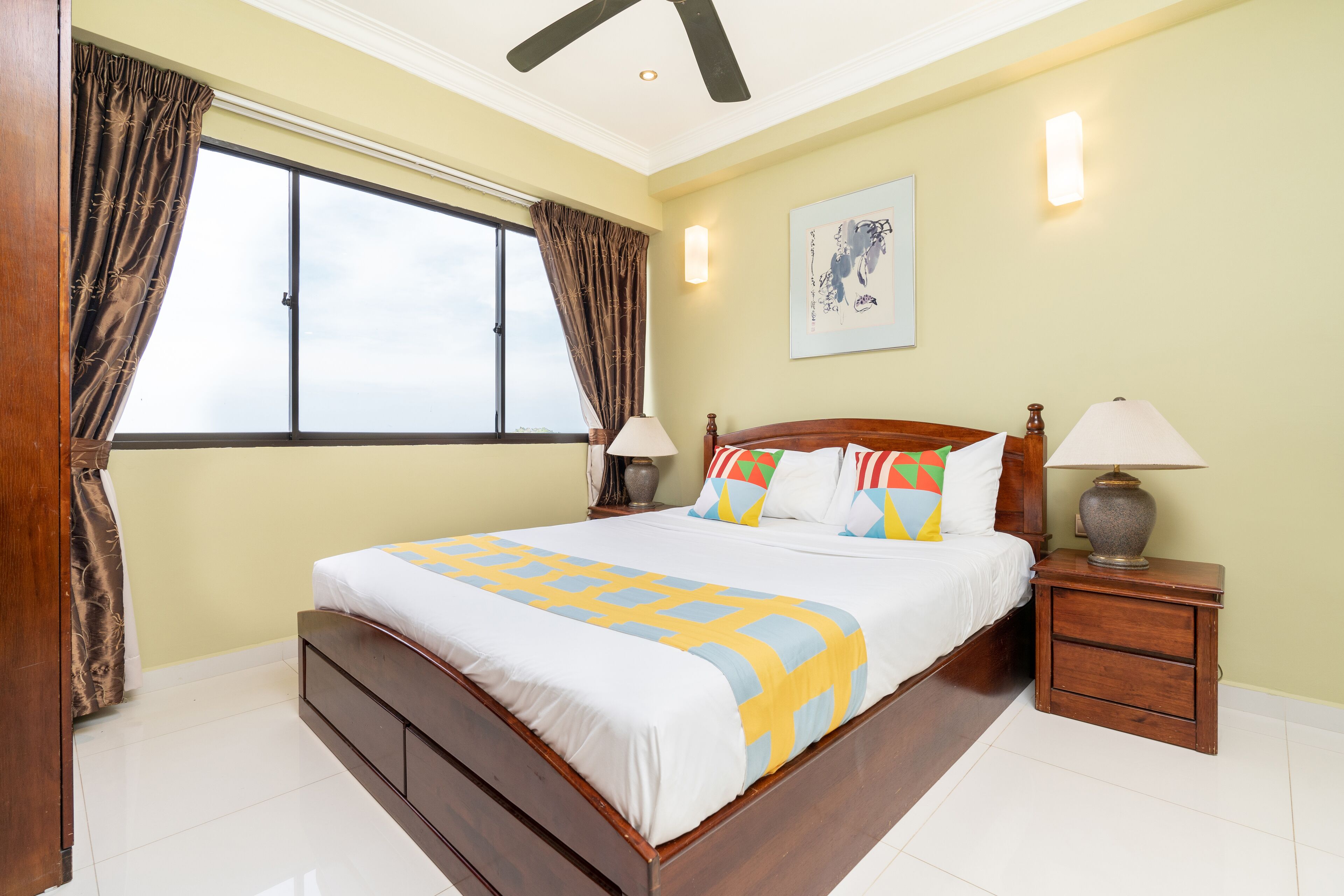 A bedroom with a large bed, two bedside tables with lamps, a window with curtains, and a wall-mounted artwork.