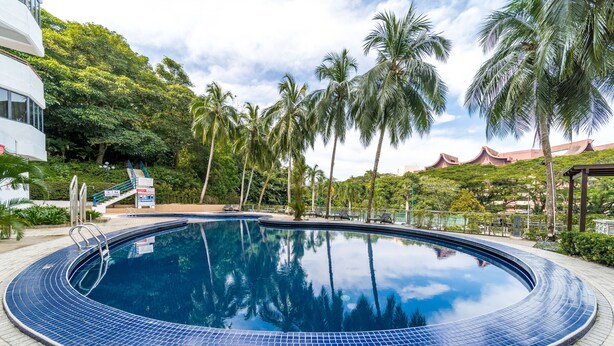 Ein Pool mit Fliesenrand, umgeben von Palmen und einem Gebäude im Hintergrund mit traditioneller Architektur.